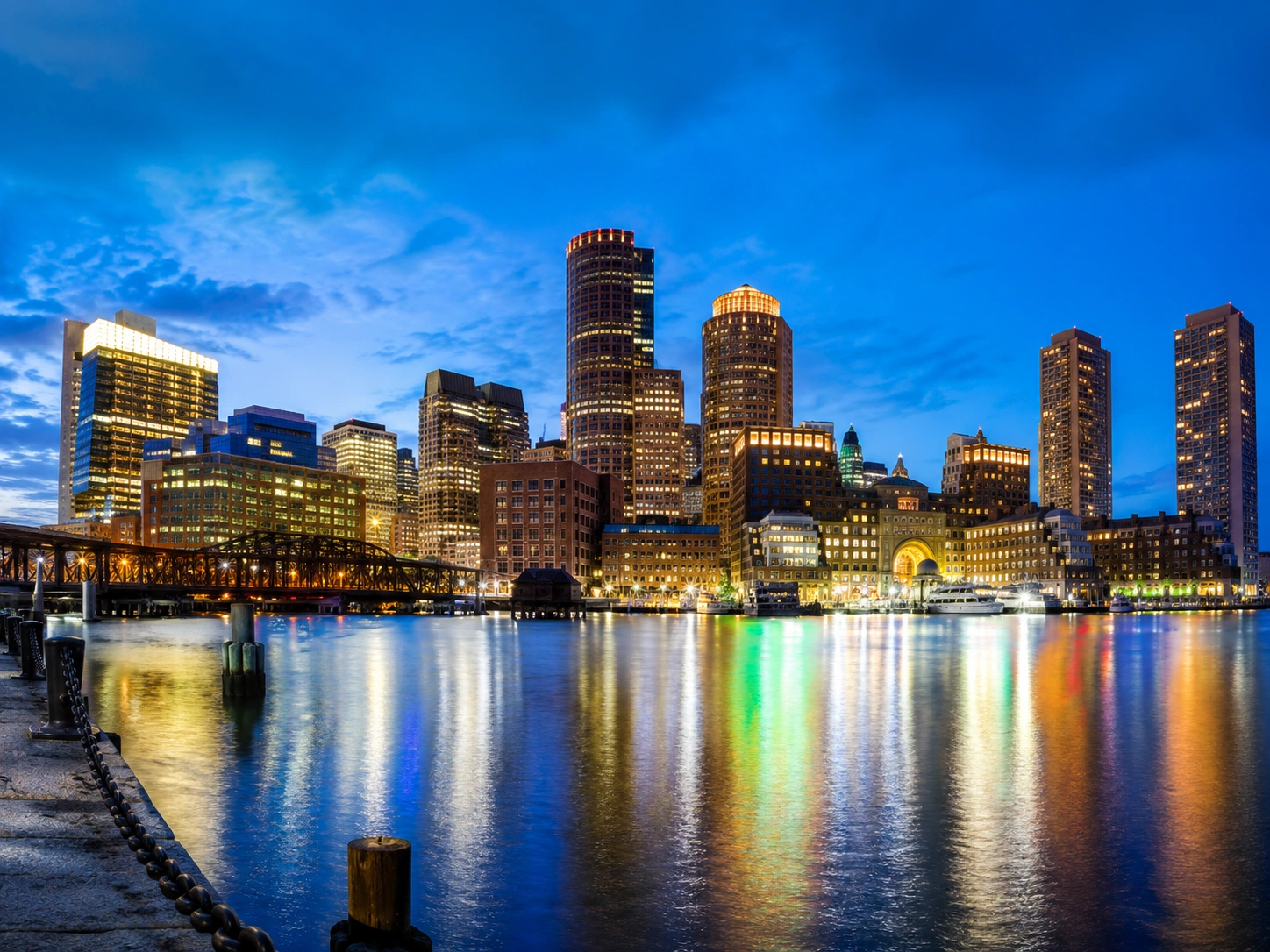 Boston skyline at dusk reflected in calm harbor waters with illuminated skyscrapers.